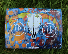 Load image into Gallery viewer, A print copy of a Capricorn star sign watercolour painting with a grey and orange background and white sea goat skull being shown off on the grass.
