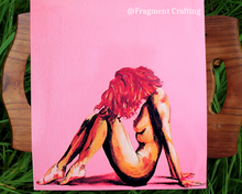 Load image into Gallery viewer, A print copy of an acrylic piece of artwork with a pink background and a nude woman sitting sideways being showcase on a board on the grass.
