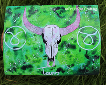 Load image into Gallery viewer, A print copy of a Taurus star sign watercolour painting with a green background and white skull of a bull being showcased on the grass.
